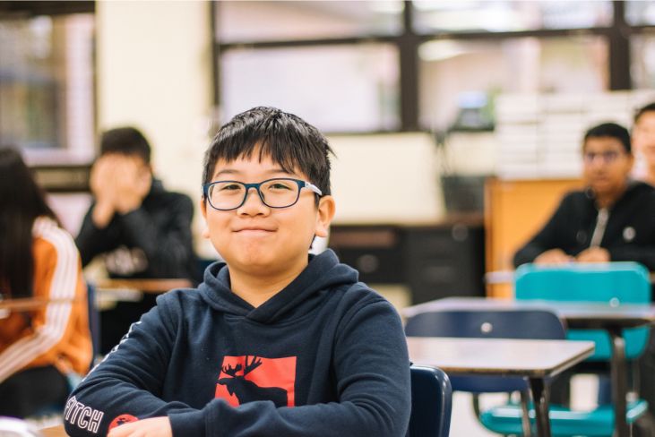 Smiling student sitting at a desk with peers in the background.